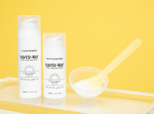 The image shows two bottles of 'The Lighter Way' body hair lightener bleach, a yellow spatula, and a clear mixing bowl, all placed on a yellow surface with a bright background.